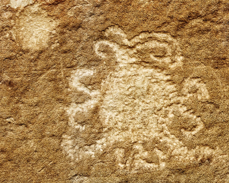 Sandstone Petroglyph in Chaco Canyon. The ancient petroglyph is pecked into a reddish rock wall. The central disk has 
			curlicues extending outward from the edge of the disk in all directions, making its diameter double of that of the central disk.  
			To the upper left of that, there is a smaller disk pecked in a similar style.